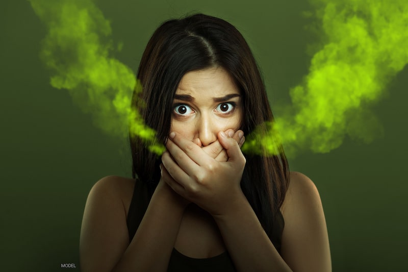 A woman covers her mouth as green smoke exits, indicating bad breath.