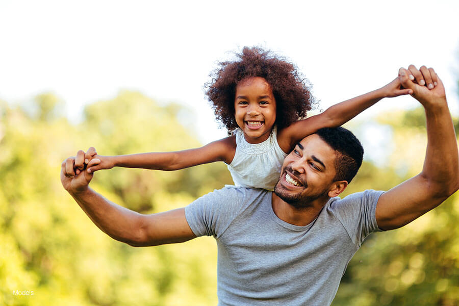 Father with young daughter on his back, both smiling with trees/green background